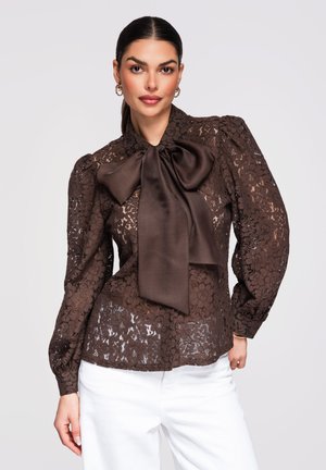 Vrouw draagt donkerbruine blouse van bloemenkant met grote satijnen strik en witte broek, poseert tegen een effen lichte achtergrond.