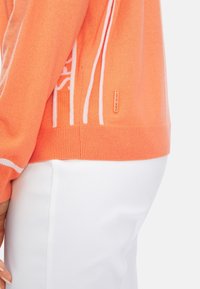 Pull orange tricoté avec des accents de design blanc, doté d'une coupe ajustée et d'un ourlet côtelé ; étiquette de marque près du bord inférieur.