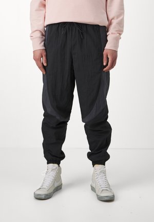 Schwarze tapered Jogginghosen mit strukturiertem Finish und elastischen Bündchen, mit grauen Akzenten und einem Kordelzug-Bund. Getragen mit weißen Sneakers.