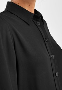 Chemise noire à boutons avec un col structuré, une texture de tissu lisse et des boutons noirs le long du plastron avant.