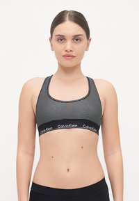 Svart og hvit stripet sports-bh med en bred elastikkant som viser Calvin Klein-logoen. Rundt utringning og racerback-design.