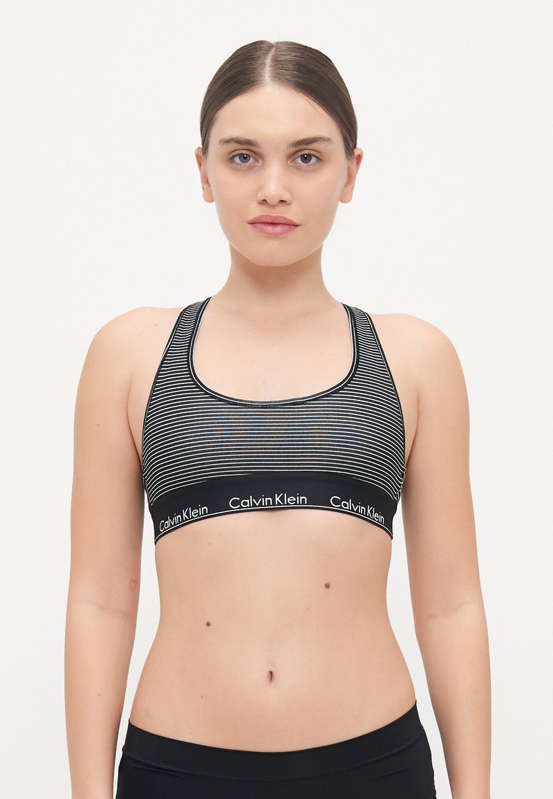 Svart og hvit stripet sports-bh med en bred elastikkant som viser Calvin Klein-logoen. Rundt utringning og racerback-design.