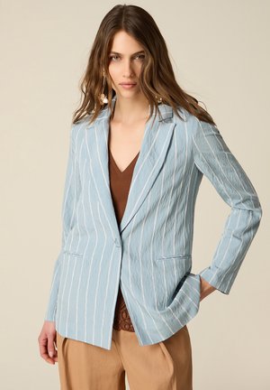 Vrouw die een lichtblauwe blazer met fijne strepen draagt over een bruin topje en beige broek, poseert met één hand in de zak tegen een neutrale achtergrond.