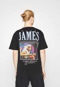 Camiseta de algodón negra con un gran texto "KING JAMES" y una gráfica colorida de un jugador de baloncesto en la parte posterior, con un eslogan debajo.