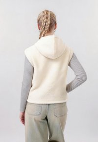 Femme aux cheveux blonds tressés portant un sweat à capuche sans manches en polaire crème par-dessus un t-shirt à manches longues côtelé gris et un jean gris clair, debout avec une main sur la hanche.