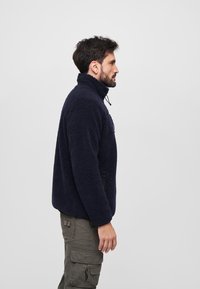 Giacca in fleece blu navy con colletto alto, tessuto testurizzato e tasche con zip, abbinata a pantaloni cargo color oliva. Vista laterale mostrata.