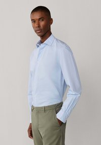 Mann trägt ein hellblaues Hemd mit Knöpfen und olivgrüne Hose, steht mit einer Hand in der Tasche vor einem einfarbigen Hintergrund.