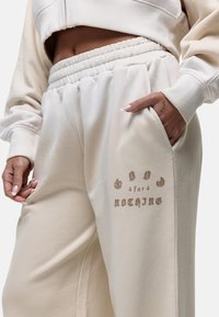 Beige cropped sweatshirt gecombineerd met bijpassende broek met een elastische tailleband en zijzak. De tekst op de broek luidt "GOD voor NIETS."