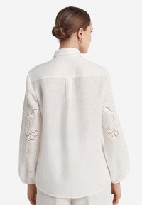 Witte linnen blouse met lange, ballonmouwen. Voorzien van fijne bloemrijke borduursels op de mouwen en een detail op de rugpas. Klassieke kraag en een comfortabele pasvorm.