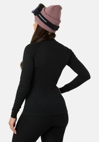 Schwarzes, geripptes Langarmoberteil mit figurbetontem Design, kombiniert mit schwarzen Leggings. Eine rosa Strickmütze wird getragen, die ein Logo und ein strukturiertes Muster aufweist.