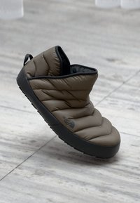 Brun polstret slip-on sko med en sort gummisål, som har en quiltet tekstur og en bageste pull-tab for nem påtagning.