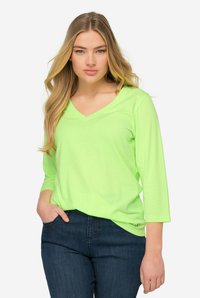 Neon-grünes V-Ausschnitt Langarmshirt aus leichtem Stoff, kombiniert mit dunkelblauen Jeans. Glatte Textur und lässige Passform.