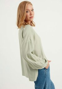 Blusa verde chiaro a maniche lunghe con vestibilità rilassata e maniche arricciate, abbinata a jeans in denim blu. Tessuto morbido, design minimalista.