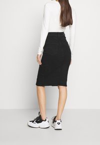 Falda lápiz de mezclilla negra con bolsillos traseros, diseño de talle alto, combinada con una blusa blanca de manga larga y zapatillas negras.