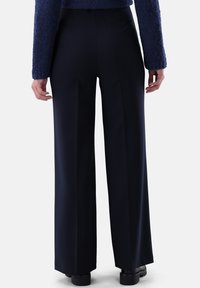 Weite Hosen aus marineblauem Stoff mit hoher Taille, glatter Textur und dezenten Falten. Von hinten mit flachen Schuhen gezeigt.