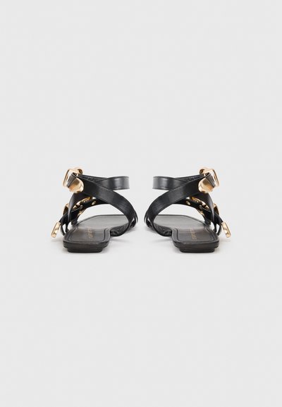 Pair of black flat sandals with crisscross straps and gold-tone buckle and rivet details, shown from the back on a white background.