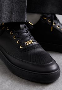 Svarta sneakers med guldfärgade öglor och märkesetikett på snöret, bärs med grå byxor och vita strumpor, på en grå golvyta.