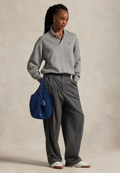 Woman wearing a grey long-sleeve polo, loose grey trousers, white sneakers, and holding a blue suede bag against a plain background.