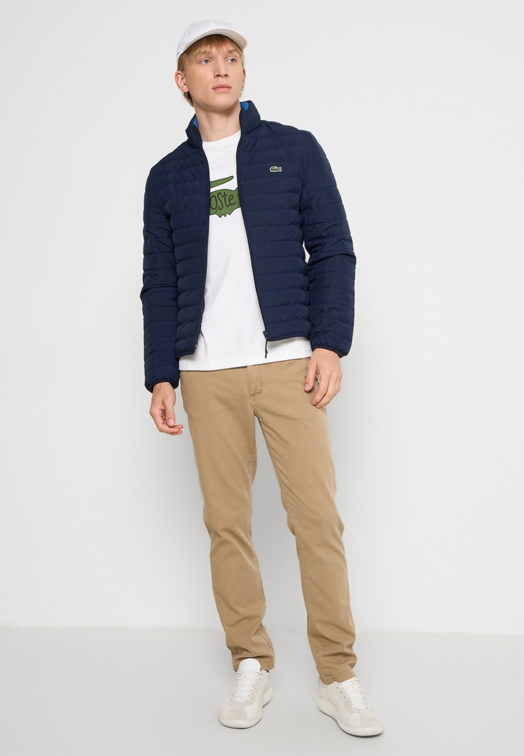 Jeune homme portant une casquette blanche, une veste matelassée marine, un t-shirt graphique blanc, un pantalon beige et des baskets blanches, adossé à un mur blanc.