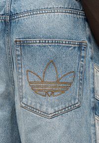 Ανοιχτό μπλε τζιν σορτς με πίσω τσέπη που φέρει καφέ κεντημένο λογότυπο. Το ύφασμα έχει ελαφρώς φθαρμένη υφή.