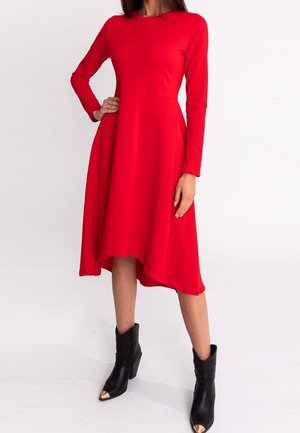 Donna che indossa un vestito rosso a maniche lunghe con orlo asimmetrico abbinato a stivaletti neri con punte metalliche, in piedi davanti a uno sfondo semplice.