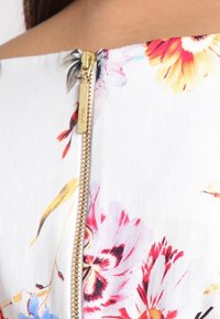 Kultainen vetoketju kukallisella valkoisella kankaalla, jossa on punaisia, keltaisia, purppuraisia ja sinisiä kukkakuvioita, näkyy vaatteen takaosassa kaula-aukolla.