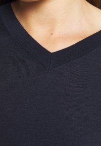 Tmavě modrý svetr s výstřihem do V, vyrobený z měkké pletené látky, s čistým zakončením na výstřihu a hladkou texturou bez viditelných vzorů.
