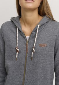 Felpa grigia con zip e cappuccio foderato, caratterizzata da lacci decorativi beige e neri, una toppa in pelle marrone con logo e una texture liscia.