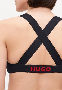 Svart sports-bh med en teksturert kant med rød "HUGO"-logo og krysset skulderstropper. Materialet ser elastisk og støttende ut.