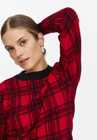 Sweter w czerwono-czarną kratę z czarnym prążkowanym dekoltem. Model ma uniesioną jedną rękę, prezentując długie rękawy swetra oraz jego tkany teksturę.