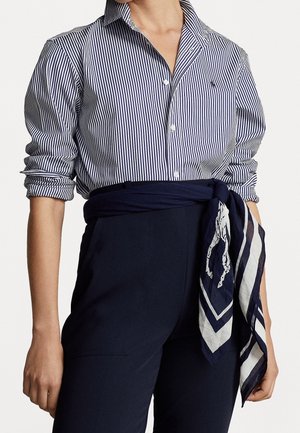 Vrouw draagt een marineblauw gestreept overhemd met opgerolde mouwen, gecombineerd met marineblauwe hoge taille broek en een gebonden marineblauwe sjaal met wit paardenontwerp.