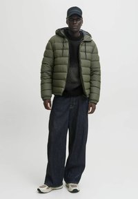 Junger Mann in einer olivgrünen Daunenjacke, dunklem Pullover, weiten dunklen Jeans, Sneakers und einer schwarzen Mütze vor einem schlichten, hellen Hintergrund.