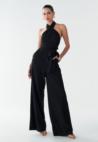 Sort halter-jumpsuit med vide ben, tekstureret stof, indsnævret talje og matchende bindebælte. Har lommer og et minimalistisk design.