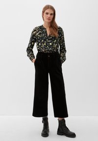 Schwarze Bluse mit Blumendruck und langen Ärmeln, kombiniert mit hoch taillierten schwarzen Cordhosen im weiten Schnitt und schwarzen Schnürkampfstiefeln.
