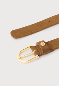 Brunt ruskindsbælte med lille guldsnørre og matchende gulddetalje med logo nær spændehullet på hvid baggrund.
