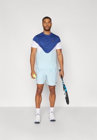 Hellblaues Sportoutfit mit einem blau-weißen, farblich abgesetzten T-Shirt und passender Shorts. Hält einen Tennisschläger und einen Tennisball.