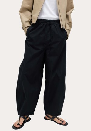 Person iført løse sorte bukser, beige jakke, hvid top og sorte flade sandaler med tåremme står på en lys baggrund.