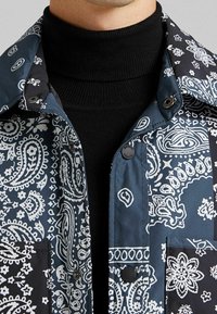 Blå och svart jacka med vita paisleymönster, med krage, tryckknappar och en texturerad svart polotröja under.