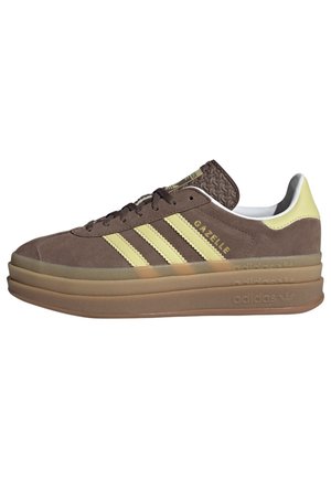 Brązowy sneaker Adidas Gazelle z żółtymi paskami, grubą warstwową gumową podeszwą i napisem "GAZELLE" po boku.