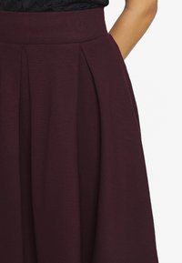Gonna plissettata bordeaux realizzata in tessuto texturizzato con vita alta e tasche laterali, caratterizzata da una superficie liscia e drappeggi delicati.