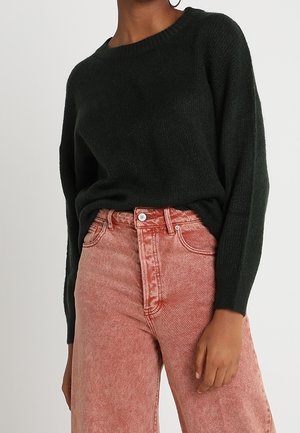 Personne portant un pull vert foncé à manches longues rentré dans un jean rose délavé taille haute, coupe décontractée.