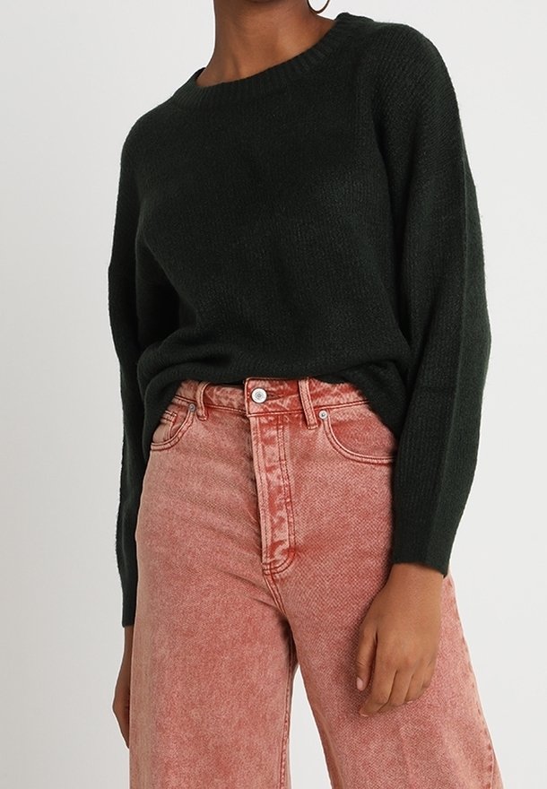 Personne portant un pull vert foncé à manches longues rentré dans un jean rose délavé taille haute, coupe décontractée.