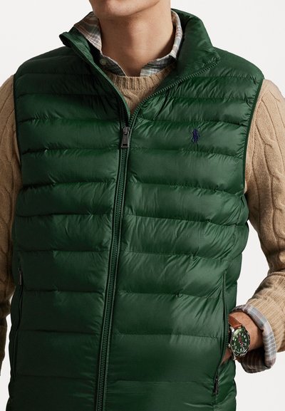 Gilet matelassé vert avec un col montant, un design matelassé, une fermeture éclair devant et un petit logo. Superposé sur un pull en tricot beige à torsades.