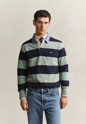 Jeune homme portant un maillot de rugby rayé vert et marine sur une chemise rose à col avec une cravate et un jean bleu, debout devant un fond uni.