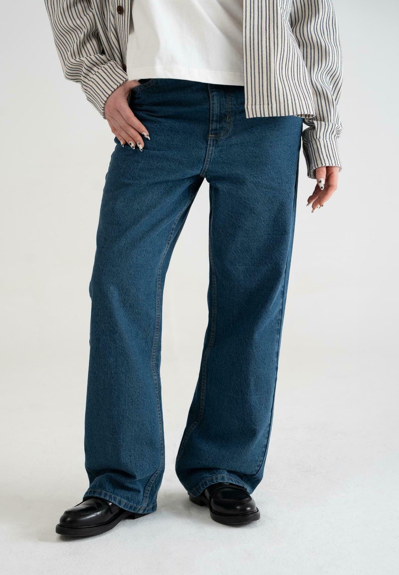 Jeans bleumarin cu picioare largi, cu o textură netedă și detalii companativoare clasice, asortați cu o cămașă cu dungi și pantofi negri.