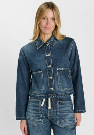 Donna con capelli biondi che indossa una giacca di denim blu scuro abbottonata e pantaloni di denim coordinati con grandi tasche anteriori.