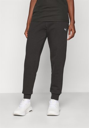 Personne portant un pantalon de survêtement Puma noir et des baskets Puma blanches, debout sur un fond uni gris clair.