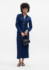 Τζιν φόρεμα με κουμπιά μπροστά, μακριά μανίκια και εφαρμοστή μέση. Σκούρο μπλε ύφασμα, συνοδευόμενο από μια μικρή μαύρη υφαντή τσάντα.