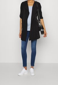 Cárdigan largo negro con frente abierto sobre una blusa azul claro, combinado con jeans ajustados azules oscuros y zapatillas blancas. Incluye un bolso negro de hombro.