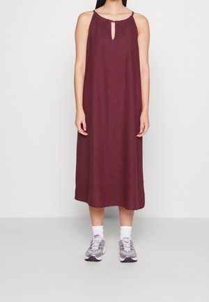 Kobieta ubrana w bezrękawnikową bordową sukienkę midi z dekoltem w kształcie klucza, białe skarpetki i srebrno-szare sportowe buty, stojąca na jednolitym tle.